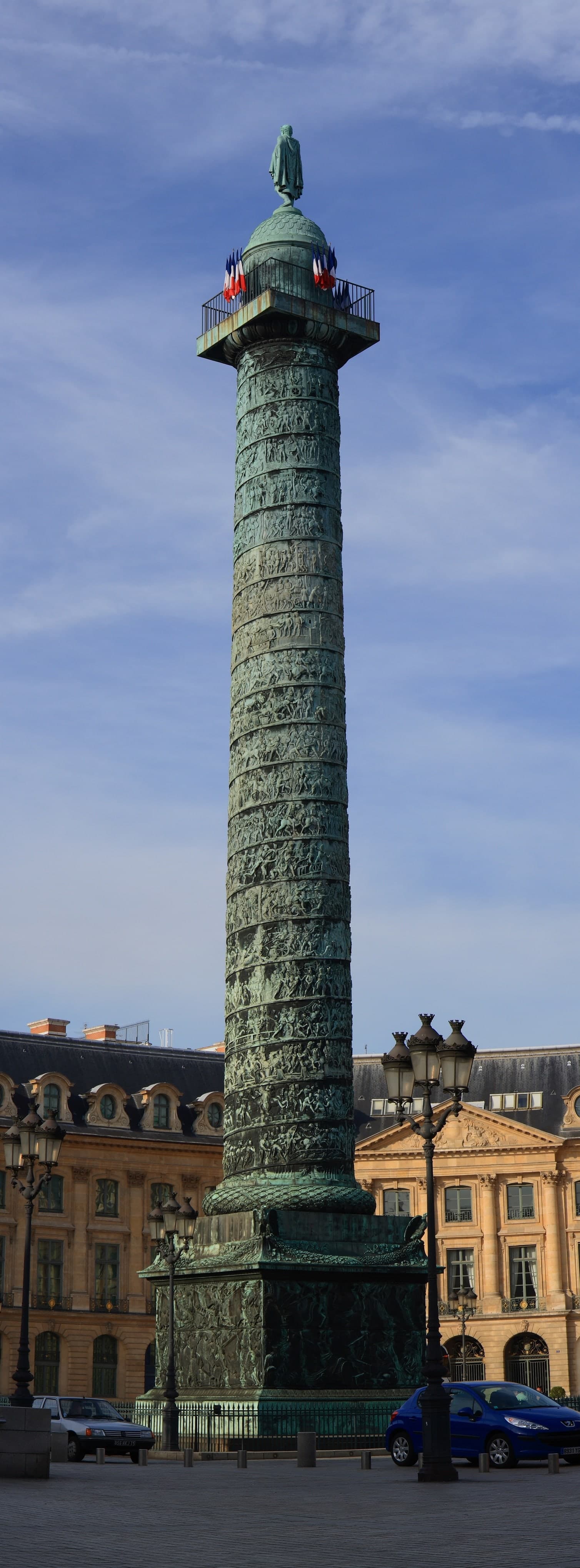 A tall slender bronze column rises at the centre of a formal square enclosed by uniform classical façades.