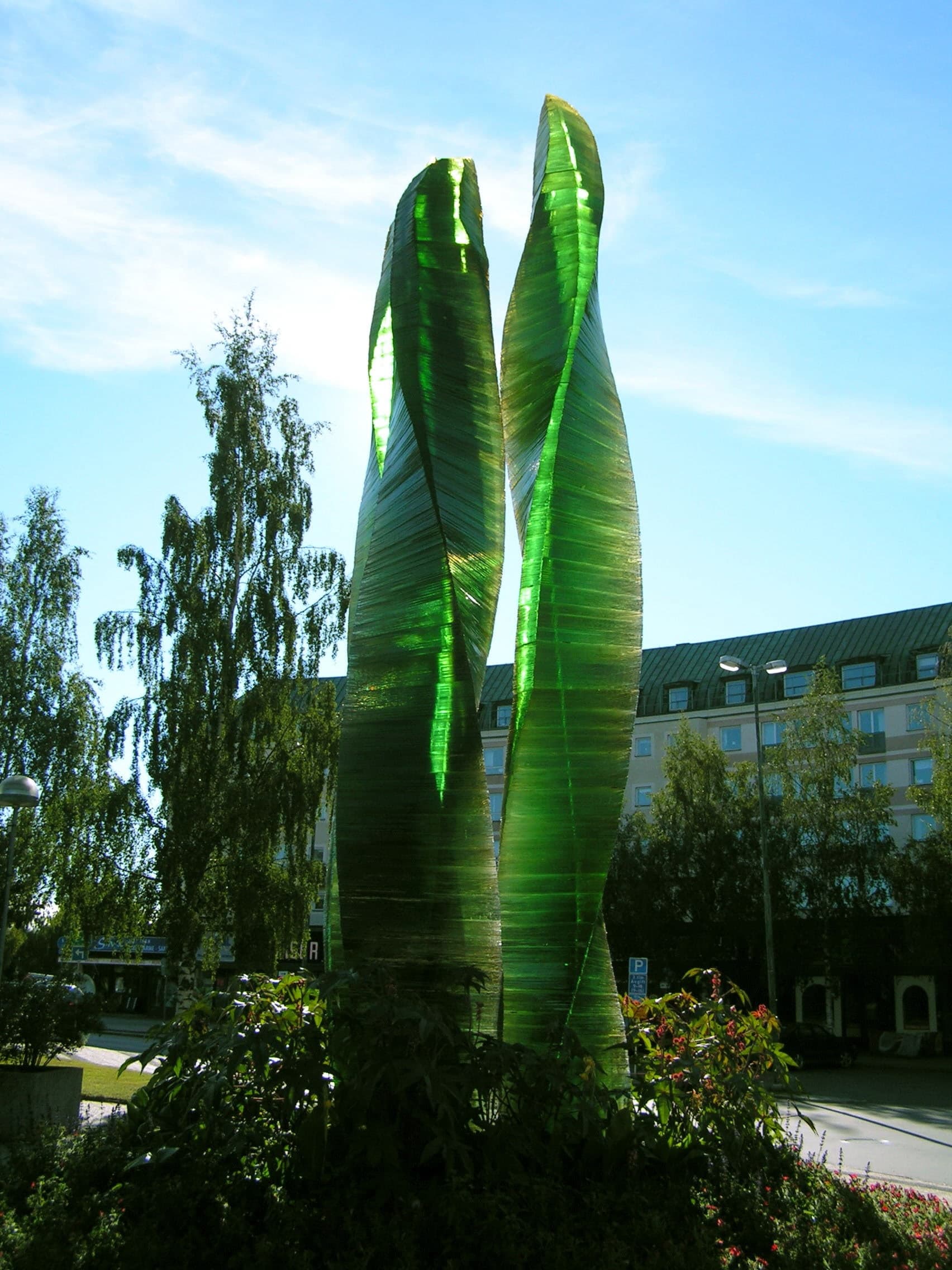 A freestanding glass sculpture in an open public square, rising clear of the paving against ordinary daylight and surrounding buildings.