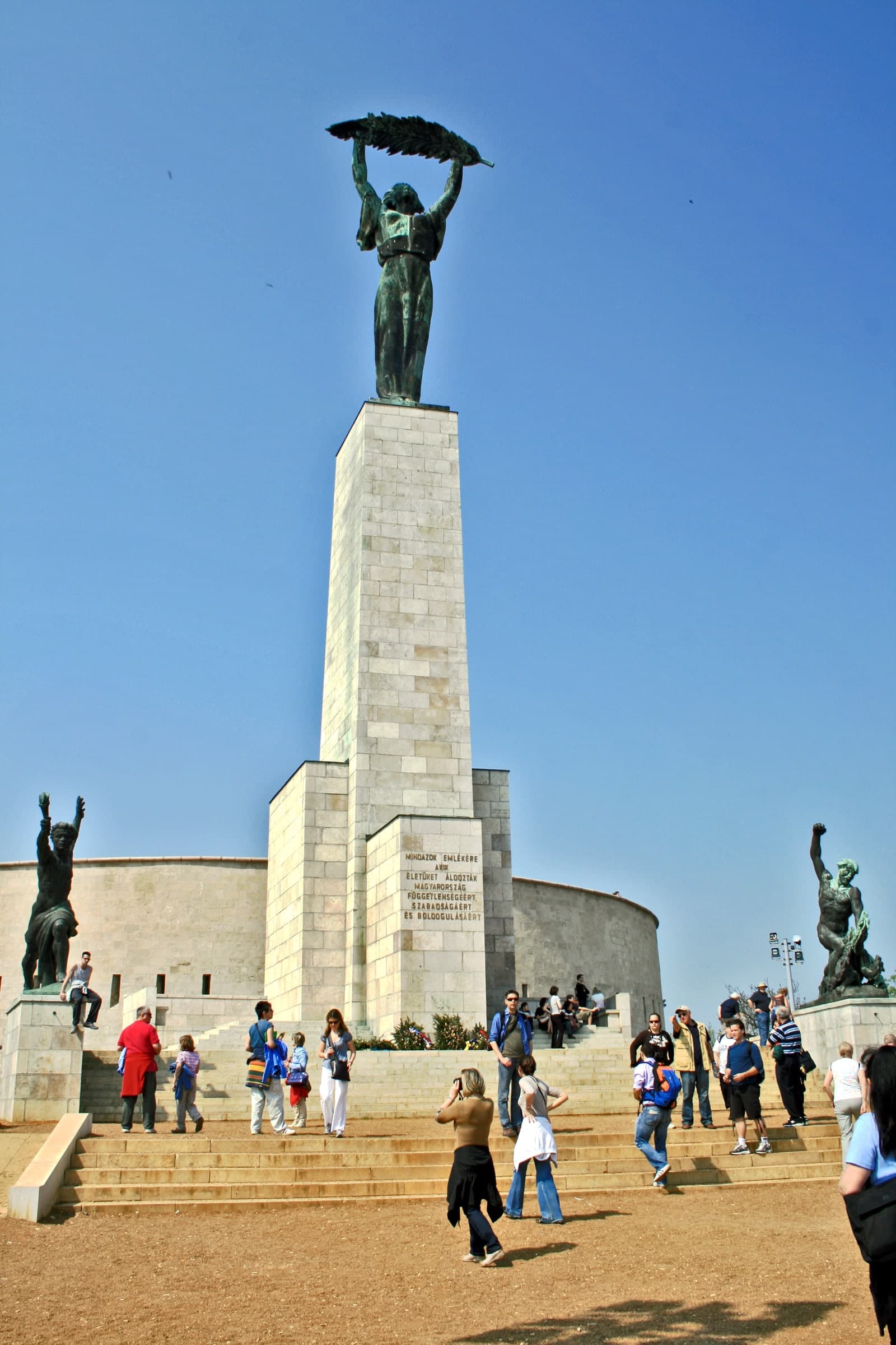 A tall commemorative monument of dark metal above a pale stone base, standing on an open hilltop ridge with the city spread below.