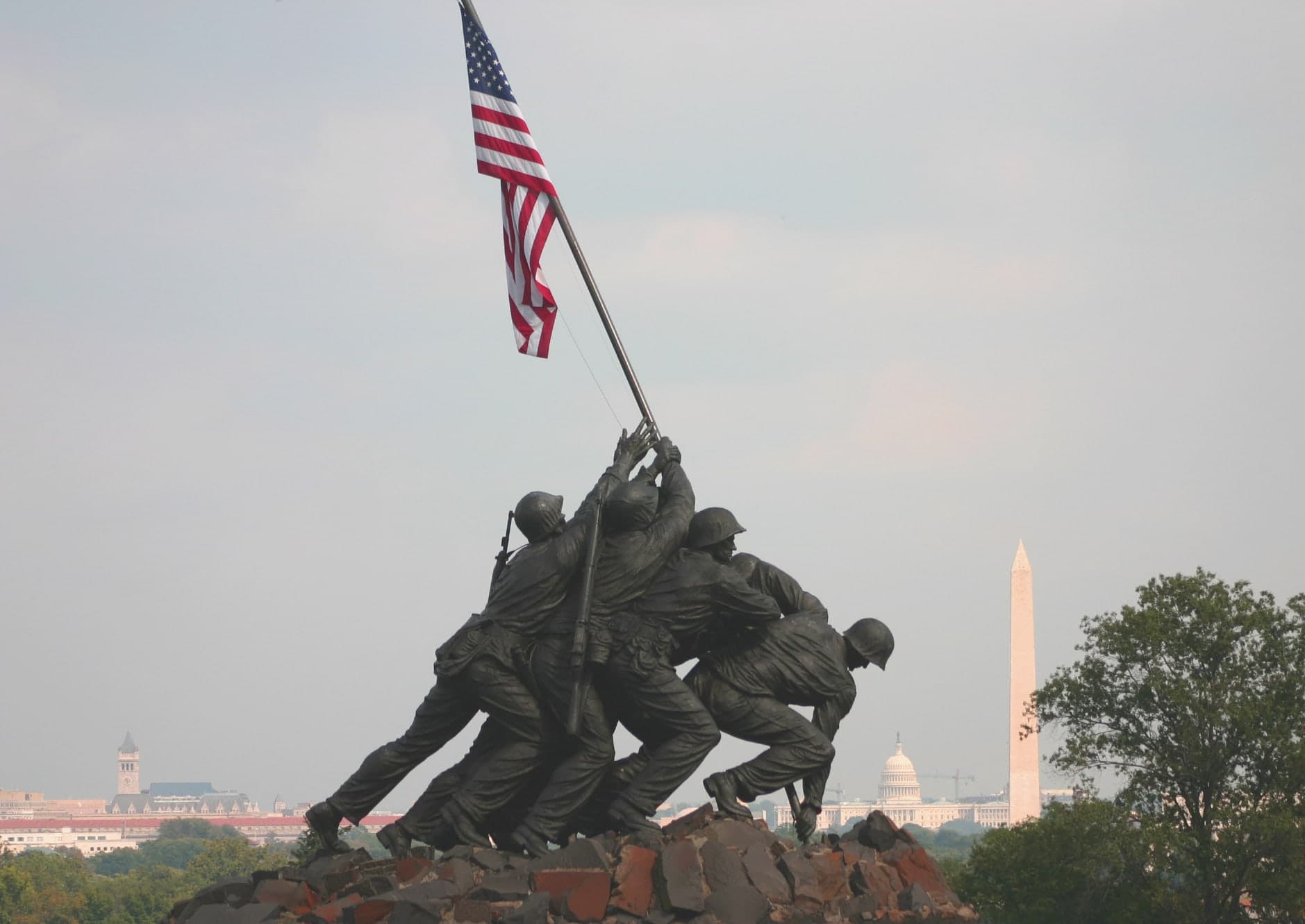 Six bronze figures cluster around a tall flagpole, bodies straining upward as a cloth flag catches the air above a broad granite and concrete base in open parkland.