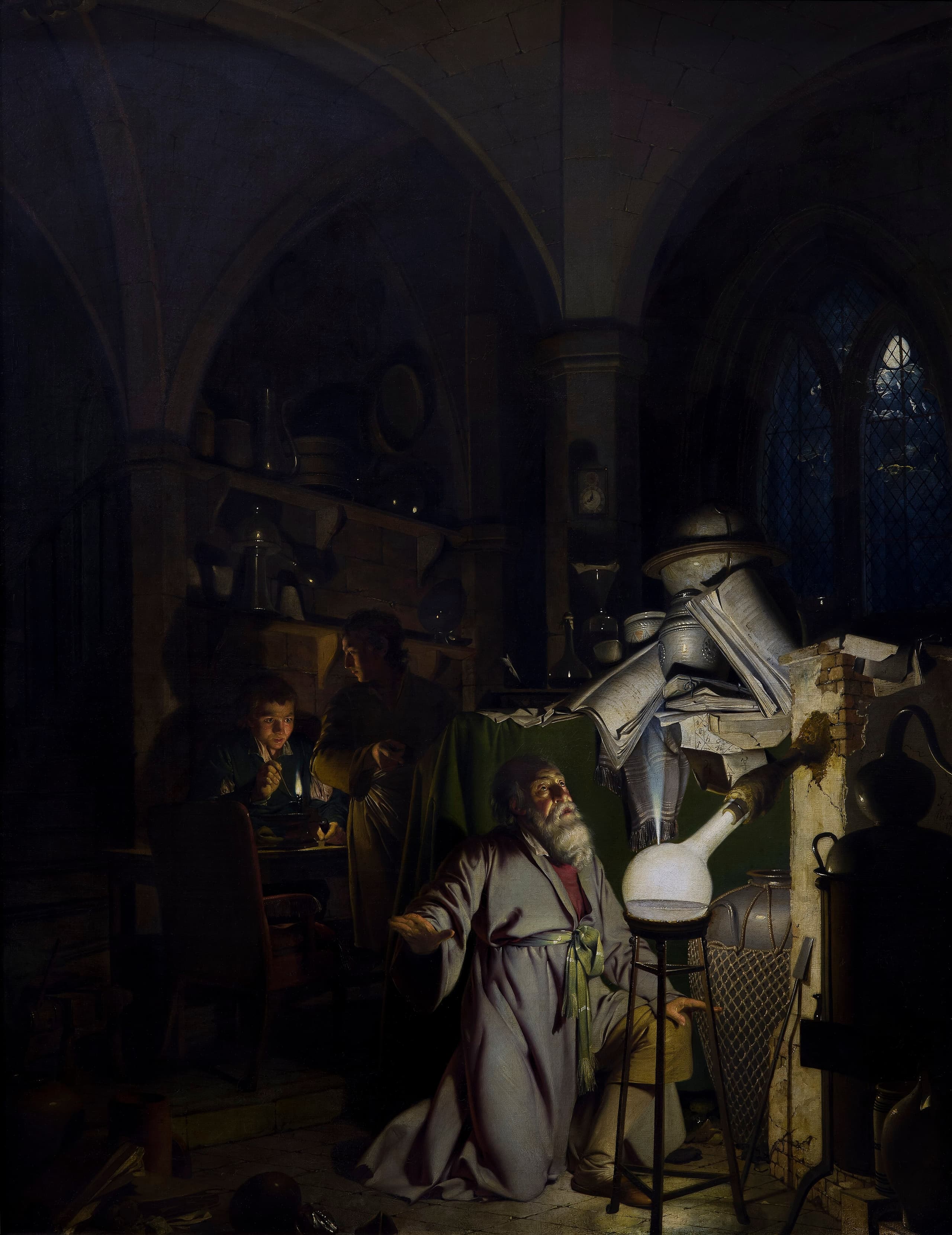 A darkened laboratory. A man kneels before glass apparatus that gives off a pale glow, hands raised toward the light. The vessel models his face and the clutter around him.