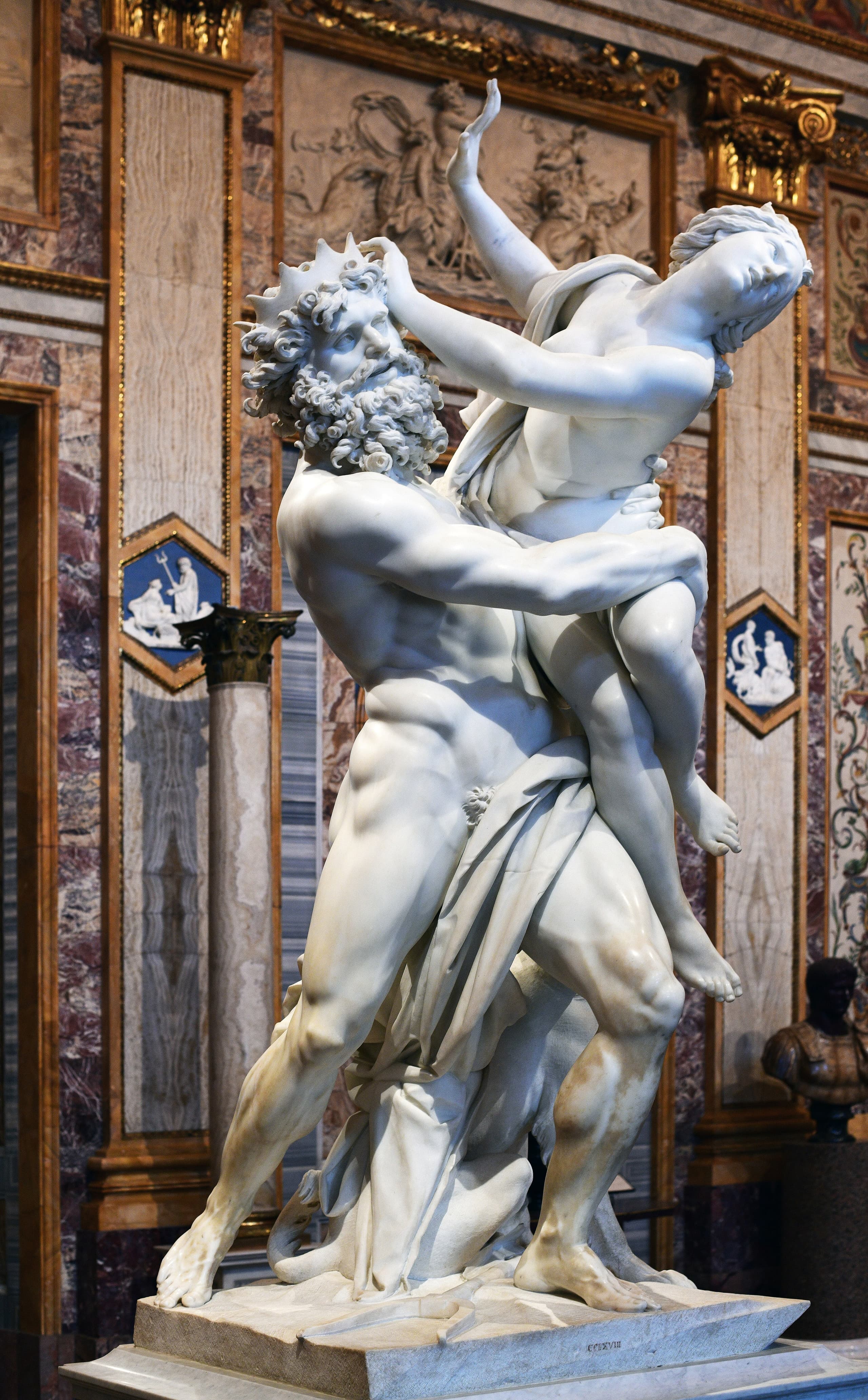 A white marble group: a muscular male figure strides as he hoists a struggling nude woman against his body; at their feet a multi-headed dog crouches, binding the pair to the base.