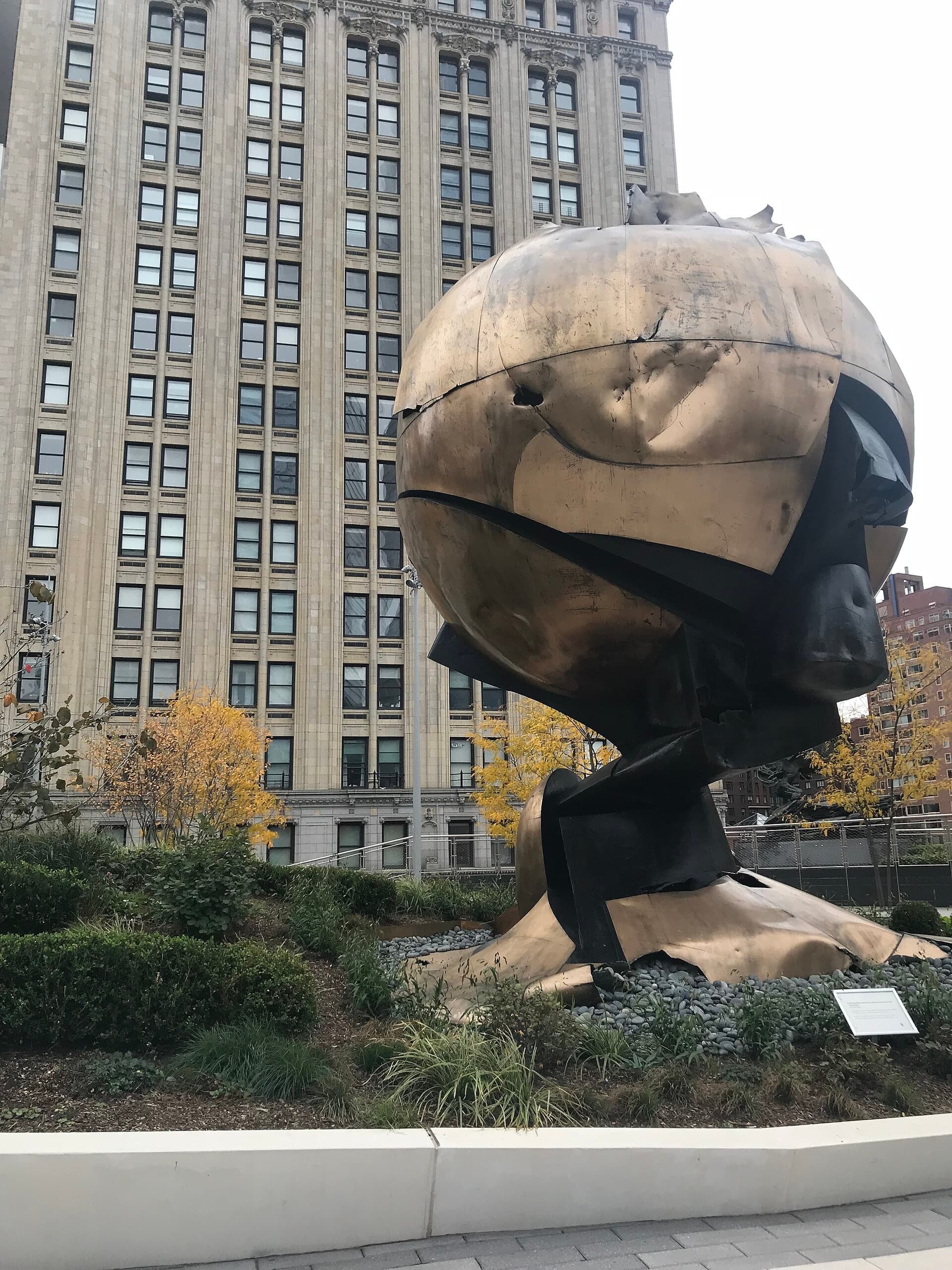 A large bronze sphere with geometric facets and openings, visibly damaged with dents and torn sections, standing on a pedestal in an urban park.