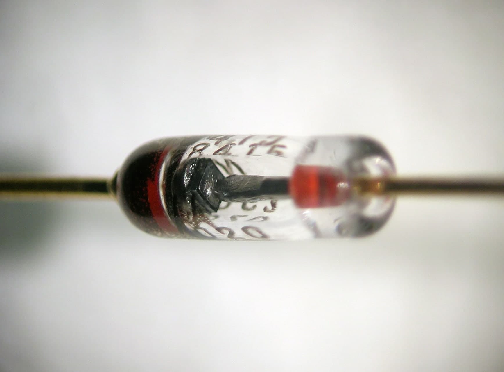 A small cylindrical electronic component with a dark body, two wire leads extending from each end, and a pale band marking one end to indicate polarity.