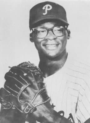 Head-and-shoulders portrait of a man in a pinstriped baseball uniform with a dark cap, looking directly at the camera, stadium seating visible behind him.
