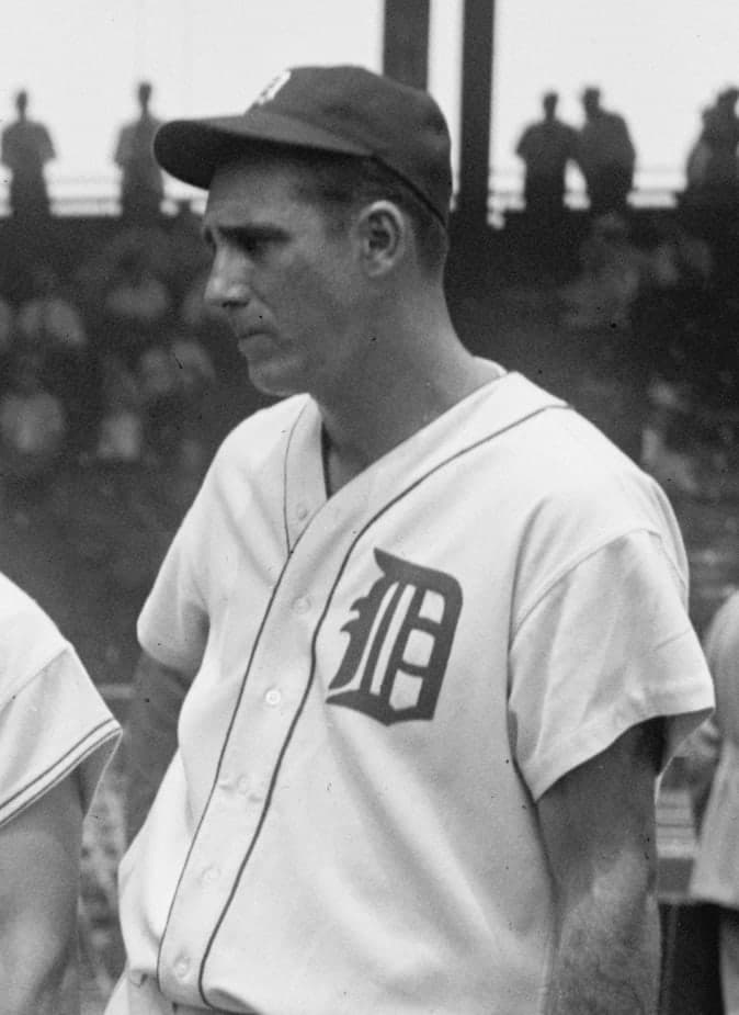 Black-and-white photograph of a tall man in a light-colored baseball uniform with a dark cap, holding a bat upright beside him, standing on a grass field with empty stadium seating visible in the background.