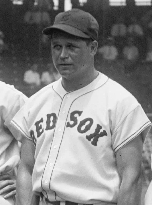 Black-and-white photograph of a baseball player in a light-colored uniform with dark sleeves, holding a bat across his shoulders and smiling at the camera.