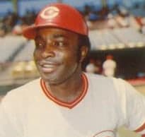 A baseball player in a white uniform with red pinstripes and a red cap, crouched in a fielding stance on a grass infield, glove extended toward the ground, stadium seating visible behind him.
