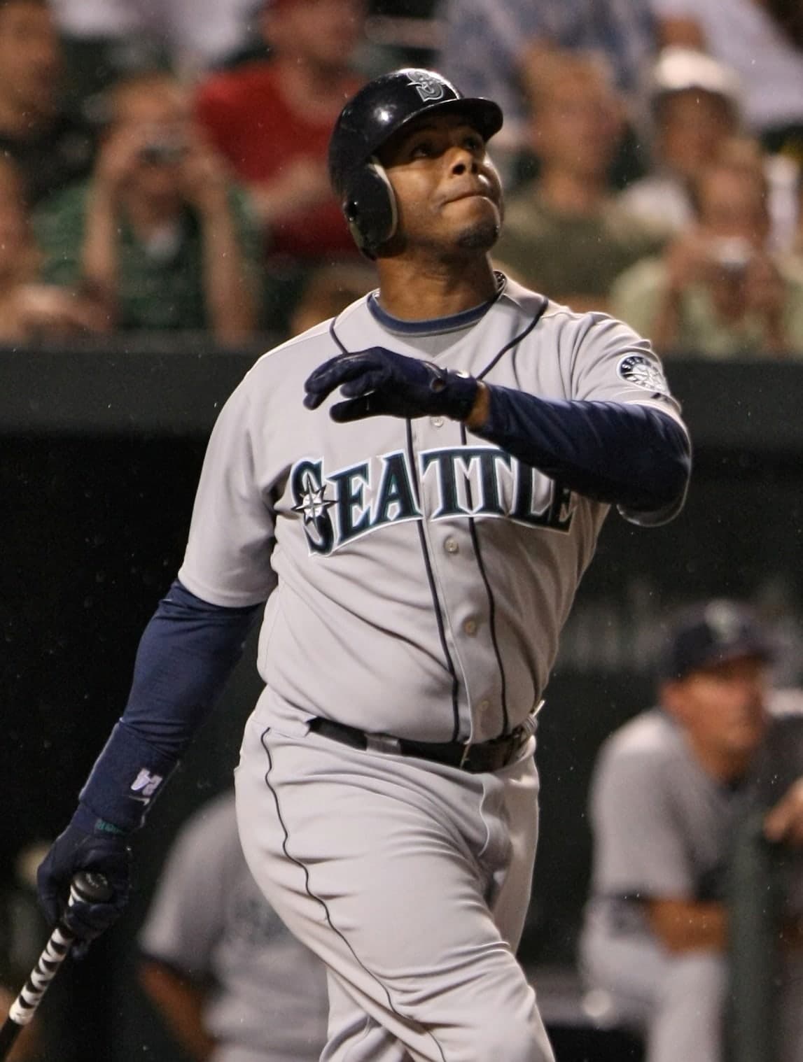 A baseball player in a dark cap and grey uniform with teal and navy piping, mid-swing, bat extended, front knee bent, back foot pivoting, with a blurred crowd in the background.