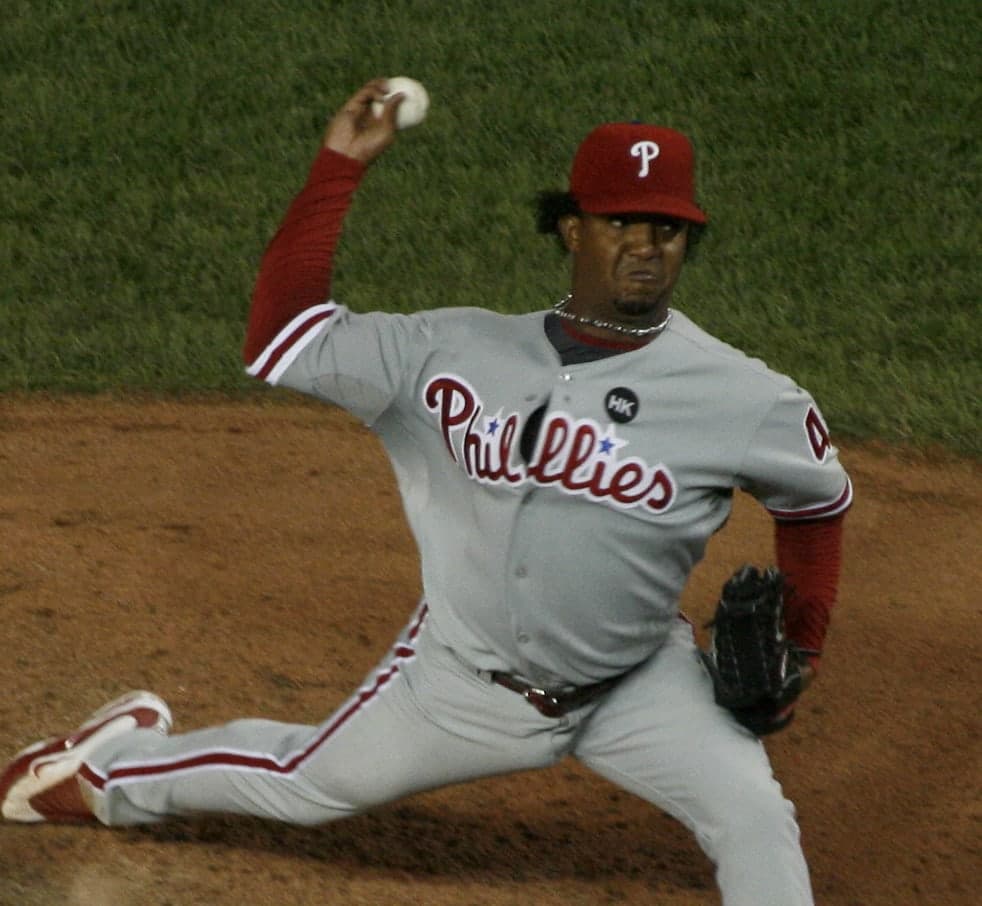 Head-and-shoulders portrait of a man in a white baseball uniform with red and blue trim, a white cap with a red letter, looking directly at the camera with a neutral expression.