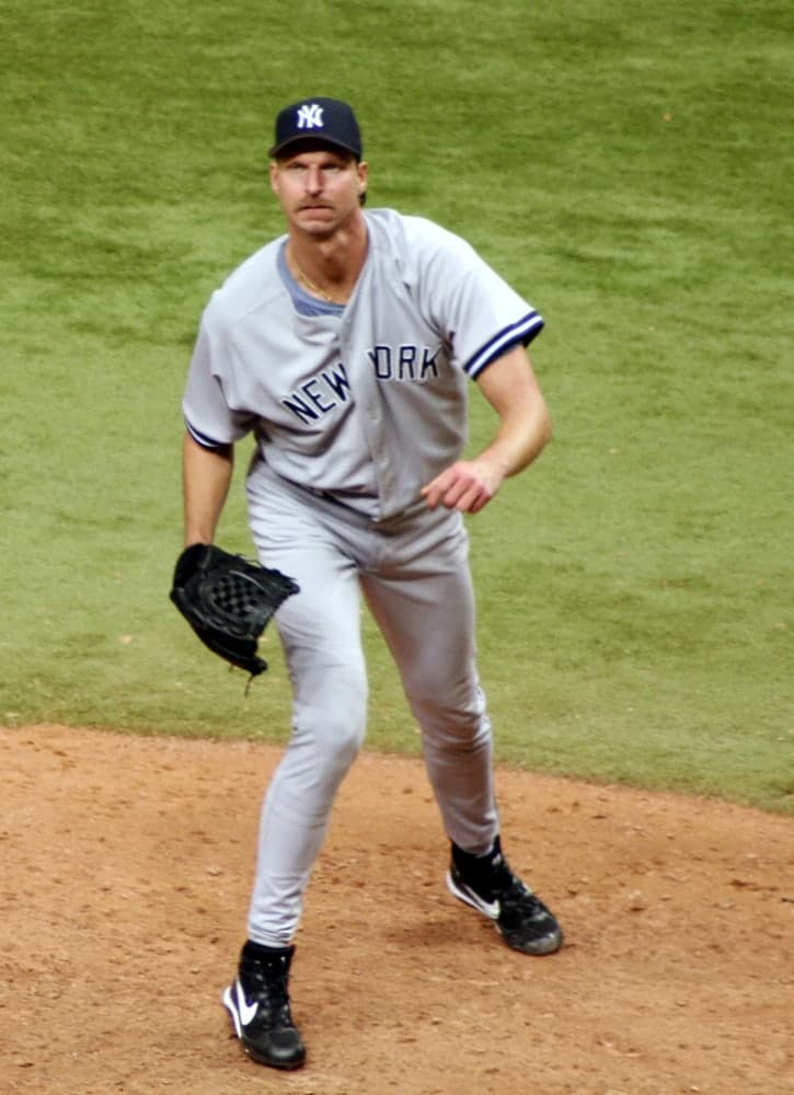 Head-and-shoulders portrait of a tall man in a white baseball uniform with purple and teal piping, a white cap with a teal bill, holding a baseball glove at chest height and looking directly at the camera.