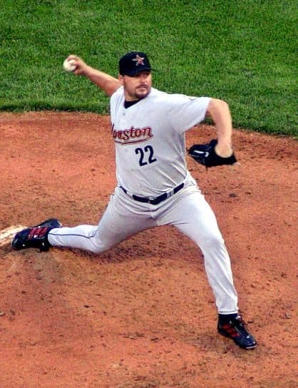 A right-handed pitcher in mid-delivery, weight shifted forward onto his front leg, left arm extended behind him for balance, wearing a white uniform with navy pinstripes and a navy cap with a white interlocking NY logo.