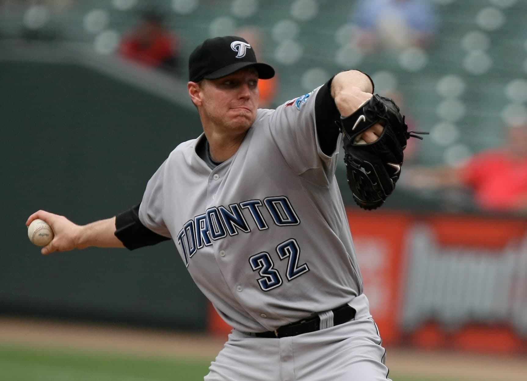 A man in a red pinstriped baseball uniform and red cap, mid-pitch, right arm extended forward and left leg lifted, on a grass field with empty stadium seating behind him.