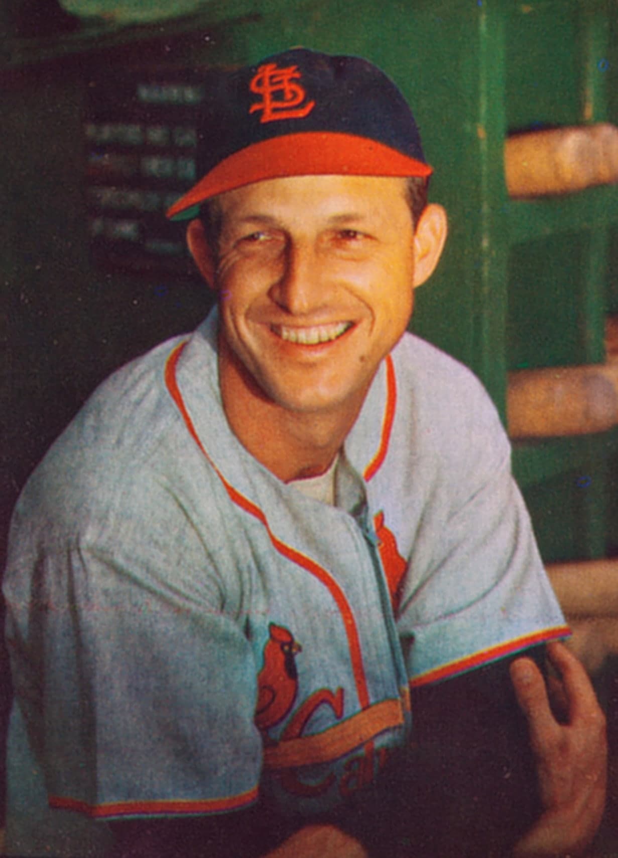 Head-and-shoulders portrait of a man in a white baseball uniform with red piping and a red cap with white letters, holding a bat over his shoulder and looking directly at the camera.
