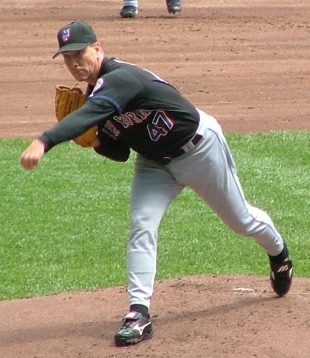 A left-handed pitcher in mid-delivery, wearing a white uniform with navy and red trim, his left leg raised high, his throwing arm extended forward, an empty stadium visible behind him.