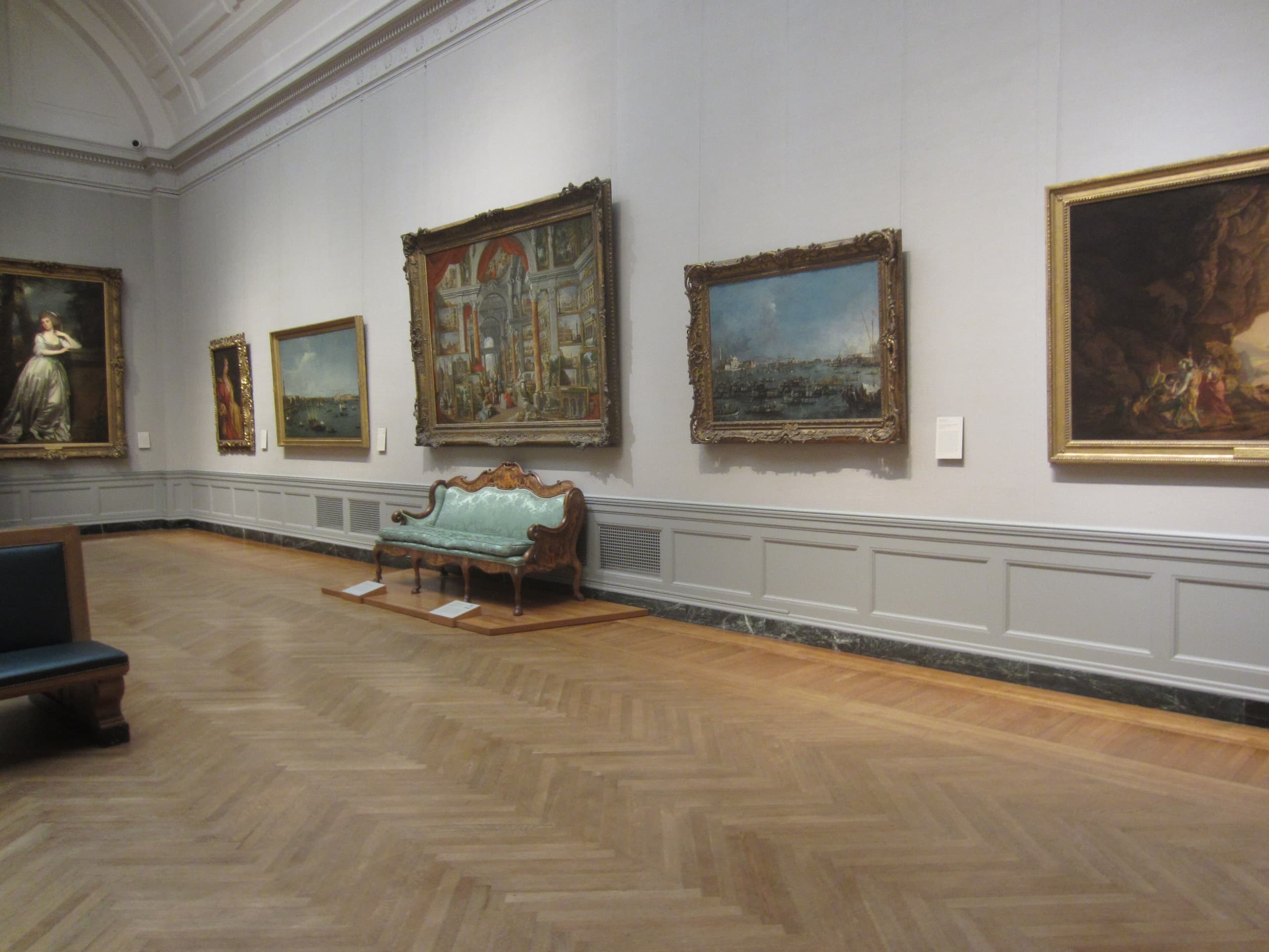 A painted gallery interior with walls densely covered in framed views of Roman buildings and monuments, with four sculptures arranged in the centre of the room.