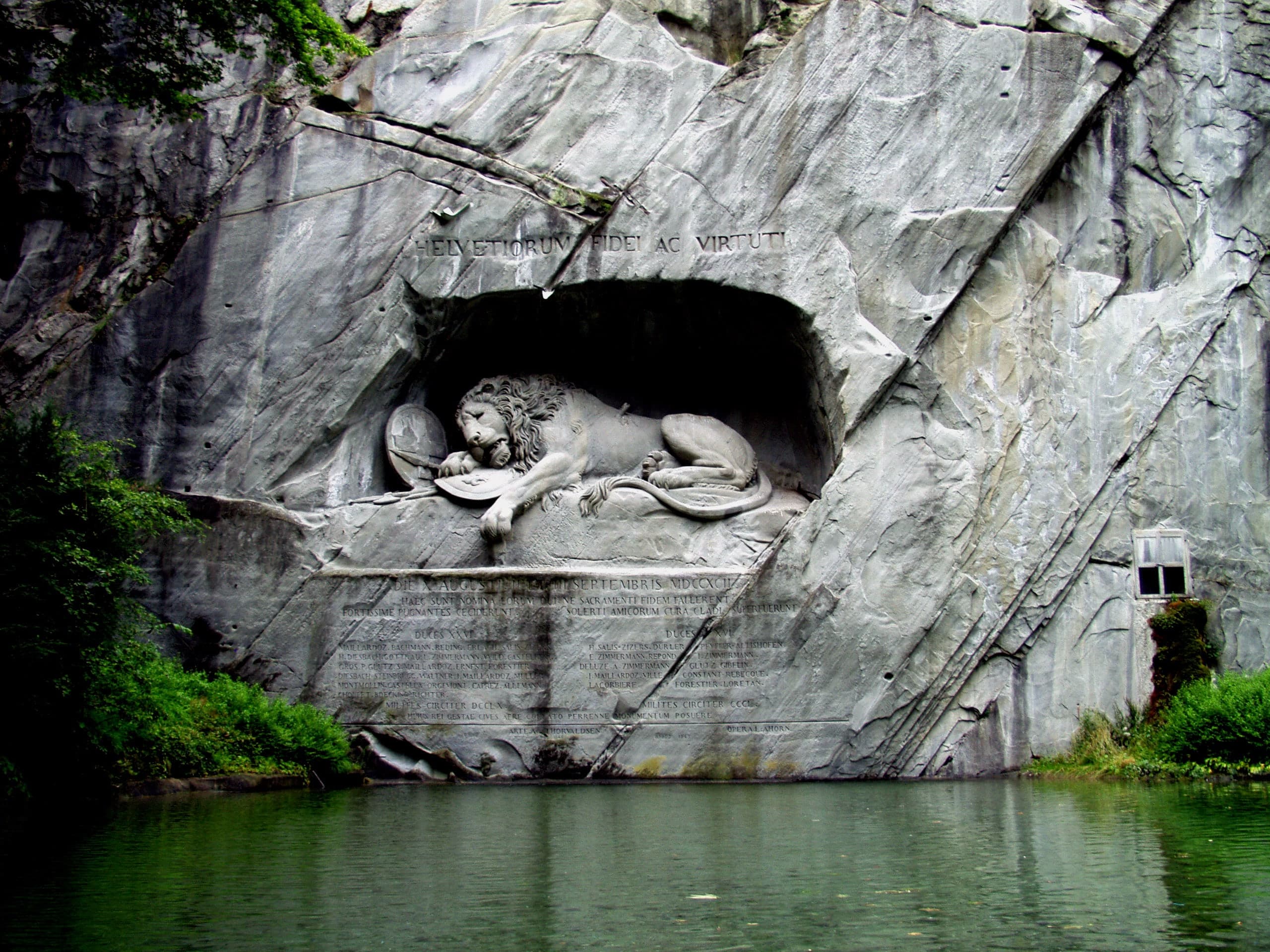 A pale sandstone rock face holds a large carved lion lying stretched and collapsed, head low, as if mortally hurt, the rough cliff continuing around the finished figure.