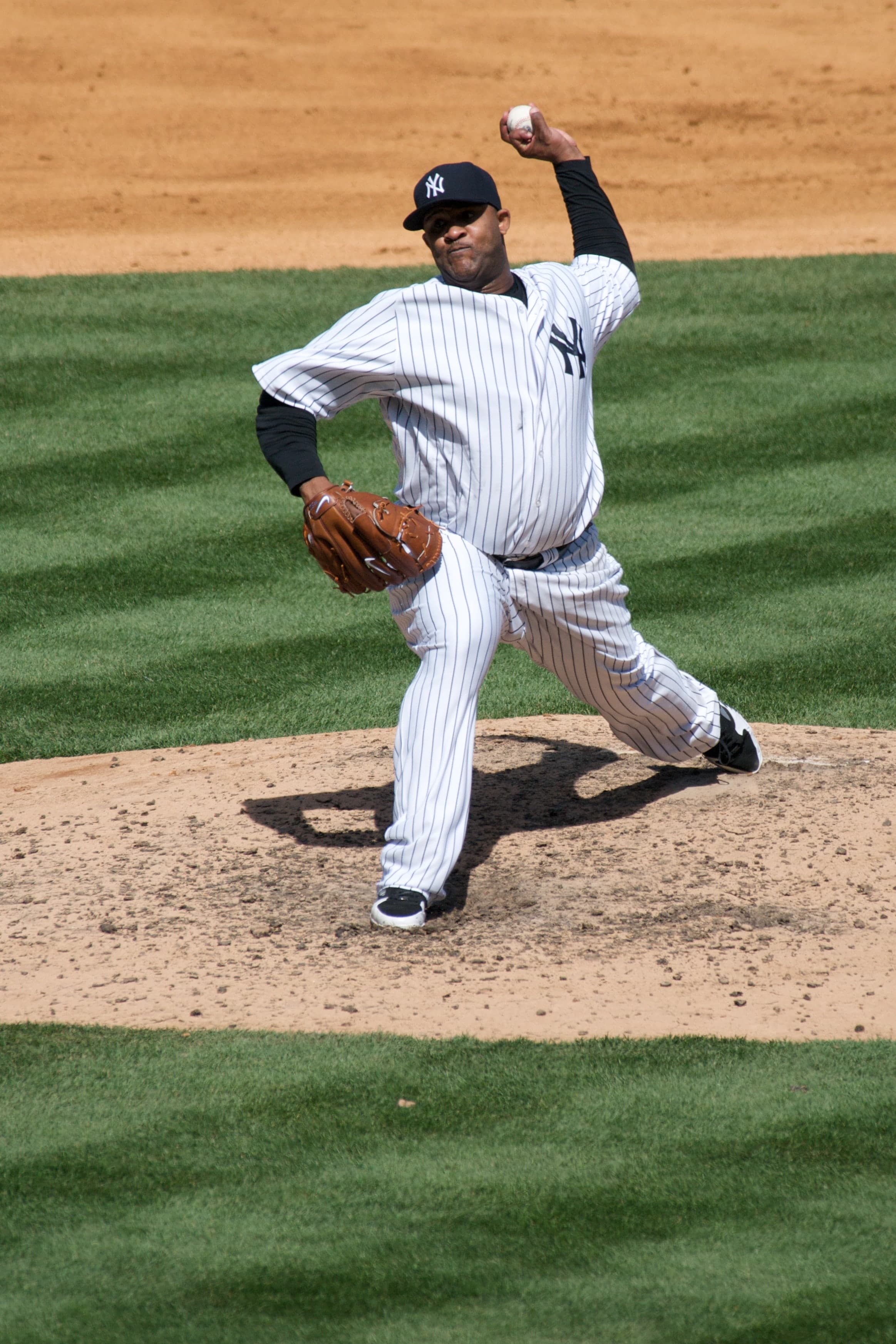 A left-handed pitcher in a grey road uniform with navy pinstripes and a navy cap, mid-delivery, right leg raised, left arm extended forward, on a grass mound in a stadium.